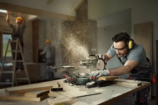 male carpenter cutting wood plank with electric saw at construction site